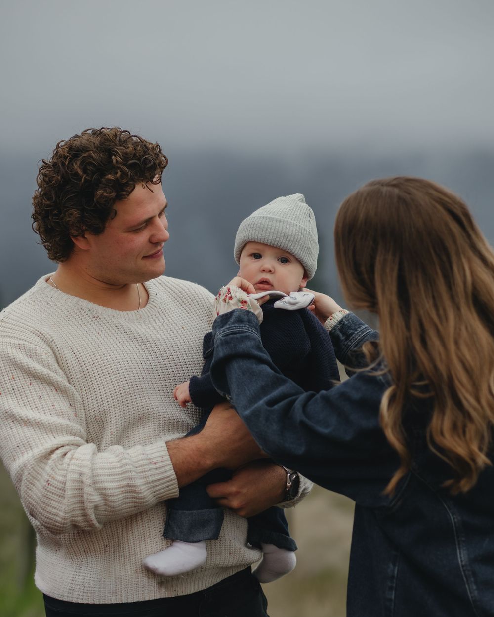 Young couple outdoors on overcast day holding their baby dressed in denim and gray beanie.