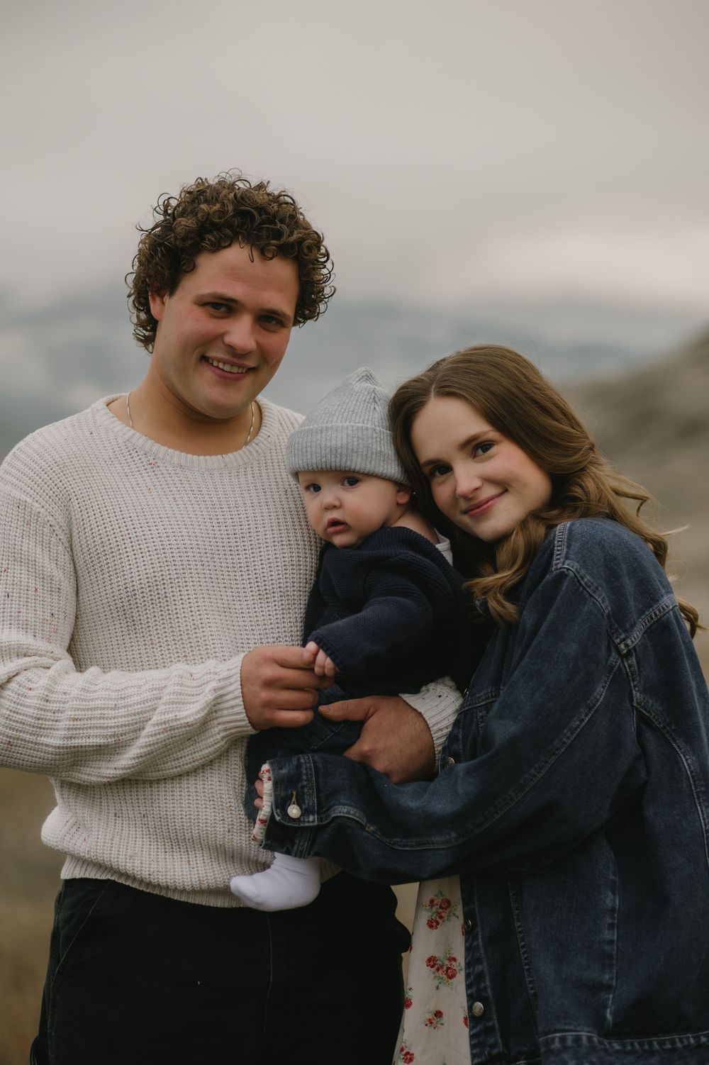 Smiling young family portrait outdoors with curly-haired father, mother, and baby in gray beanie.