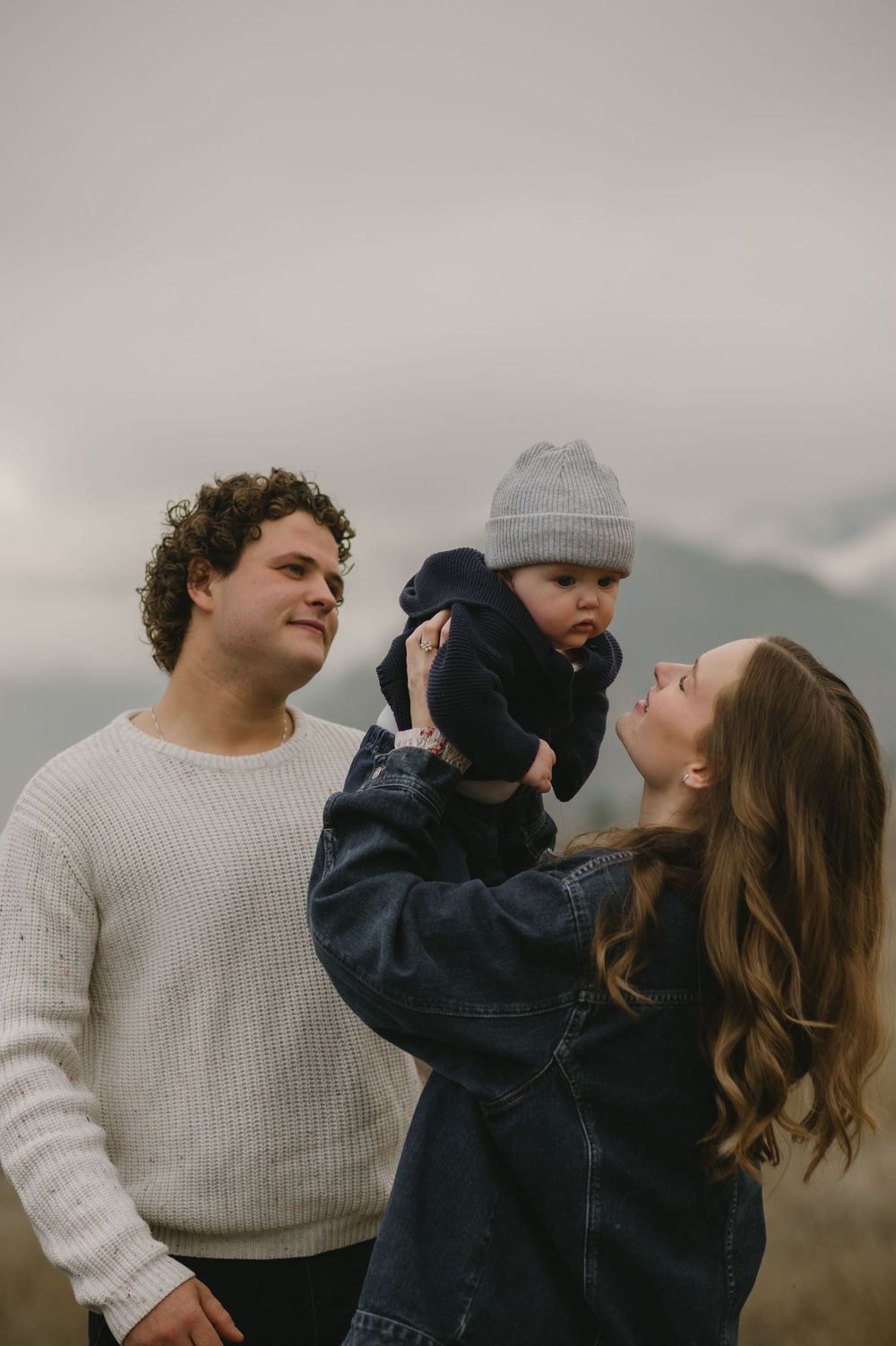 Young family outdoors, father in cream sweater smiling as mother lifts baby in gray beanie against moody sky.