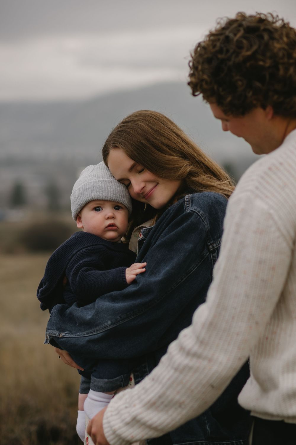 Mother in denim jacket tenderly holds baby in gray beanie while father looks on in misty outdoor setting.