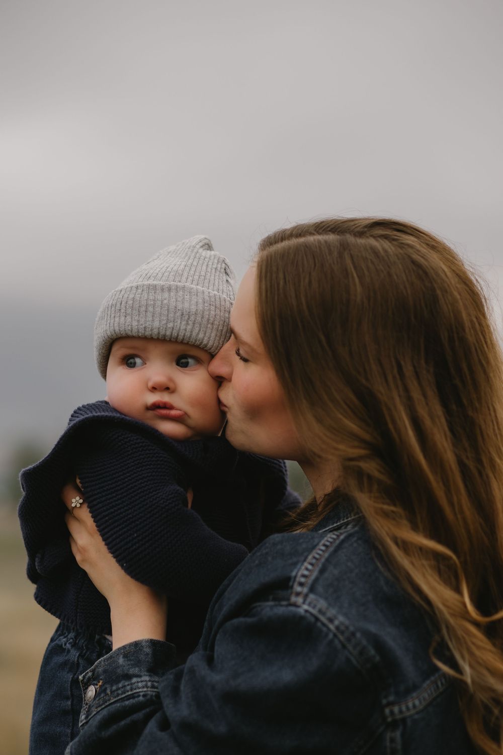 Mother gently kisses baby wearing gray knit beanie during an outdoor family photo session.