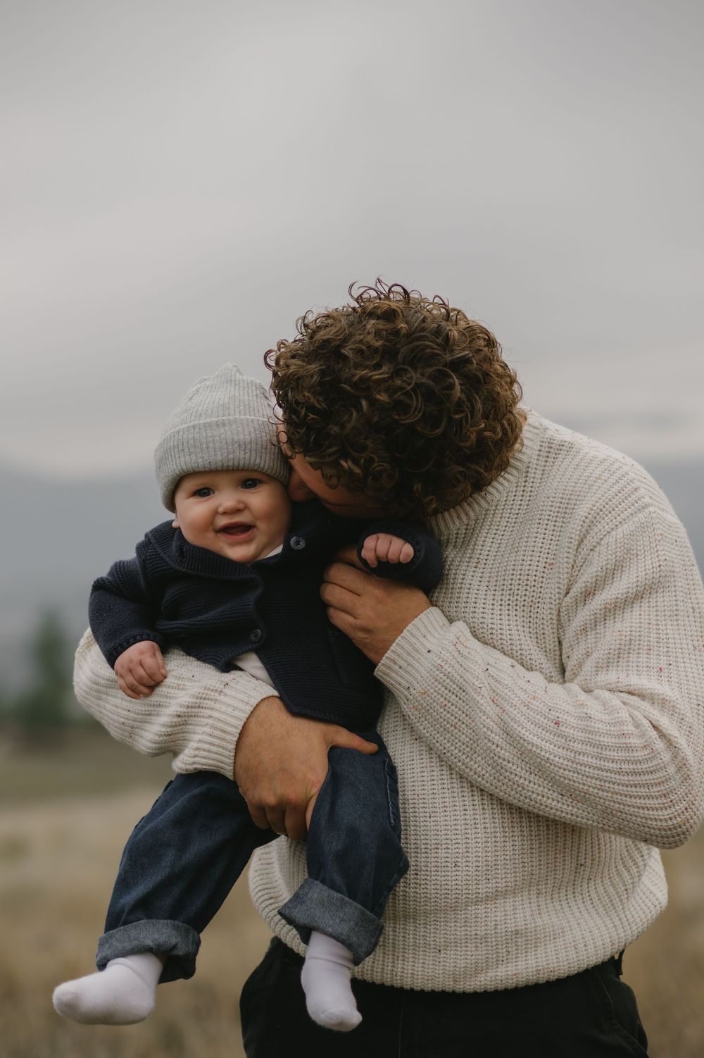 Curly-haired father in cream sweater kisses smiling baby in gray beanie during outdoor family shoot.