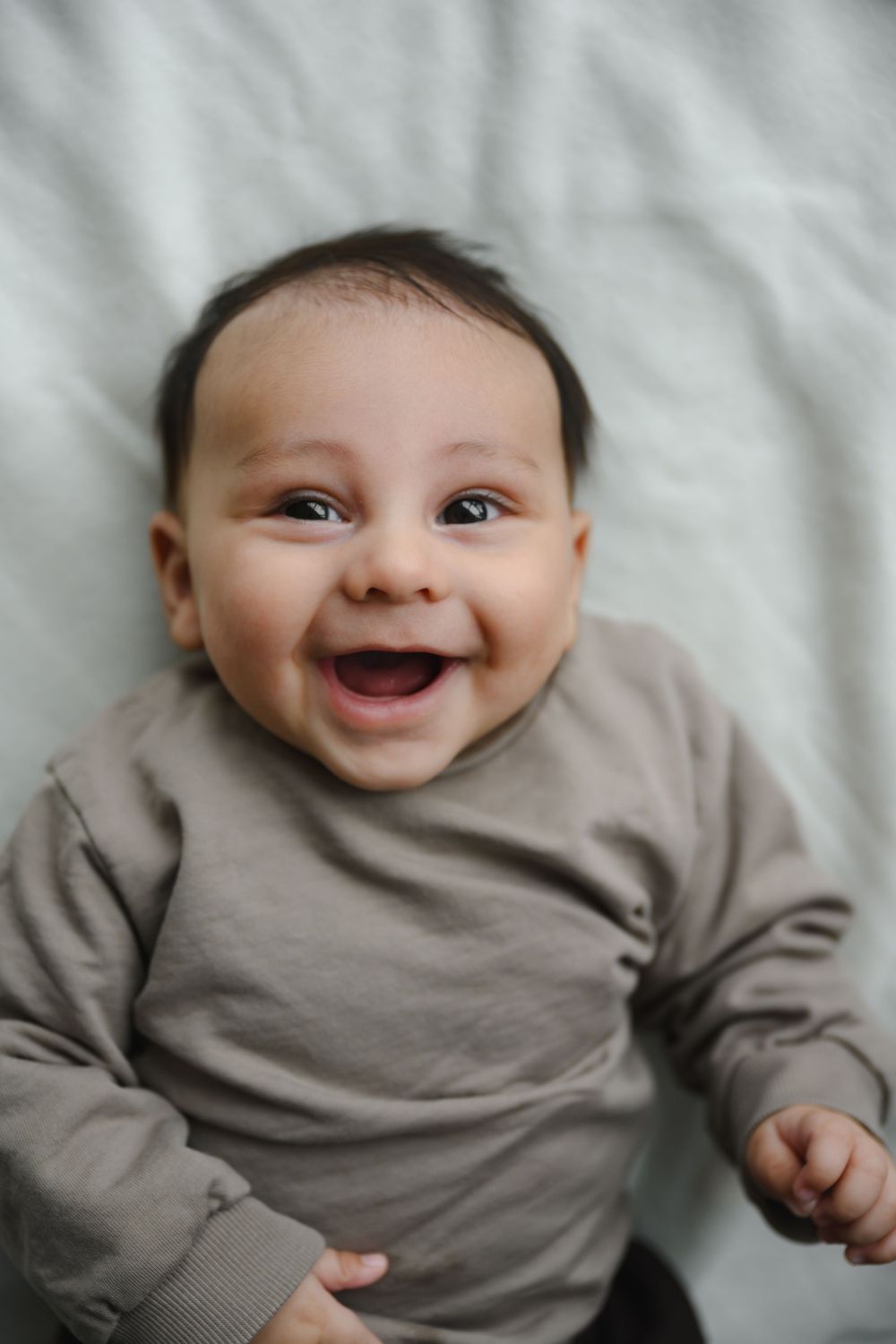 An adorable smiling baby in a gray outfit lies on a white blanket with a big happy grin.