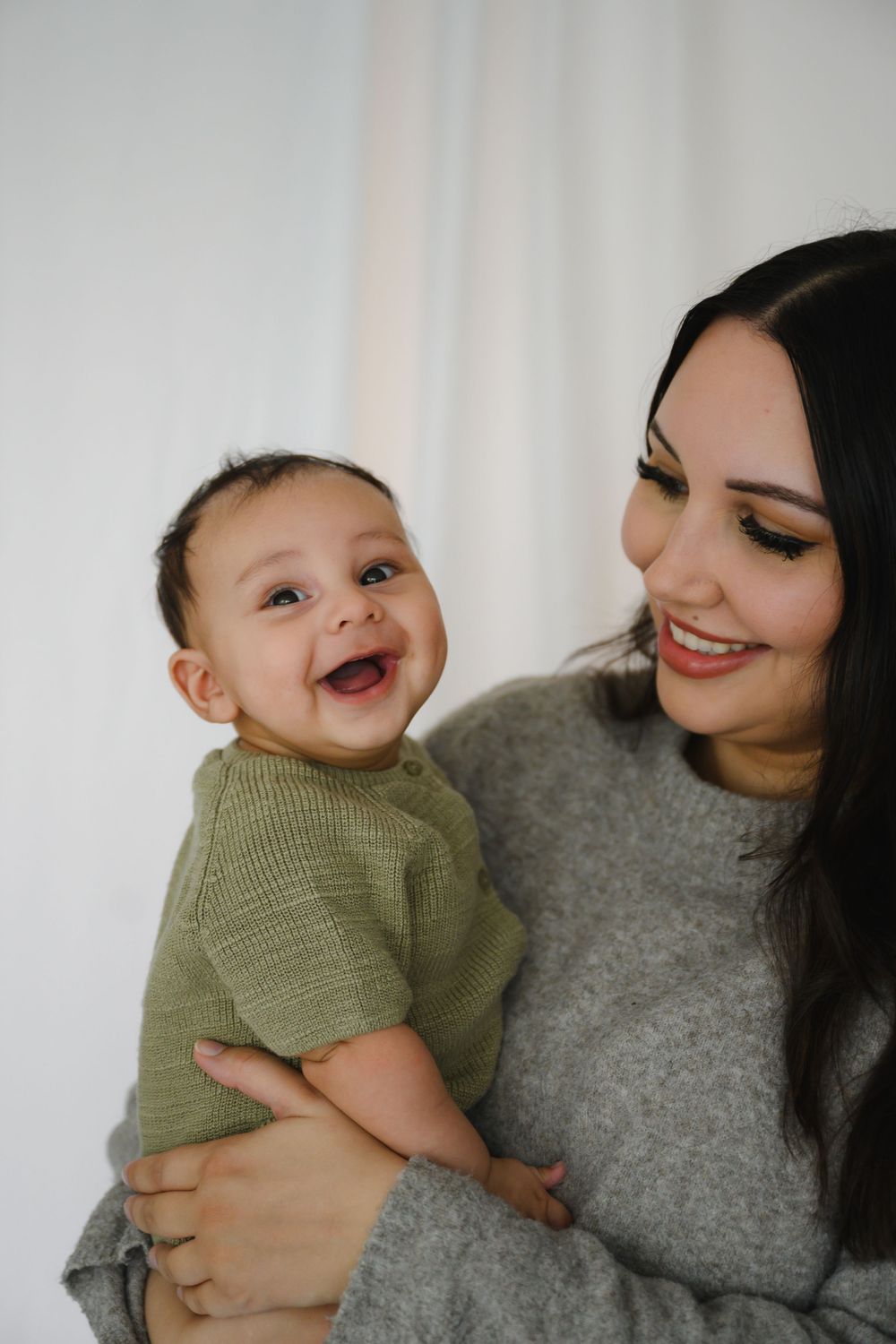 Smiling mother in gray sweater holding laughing baby in green knit outfit against white background.