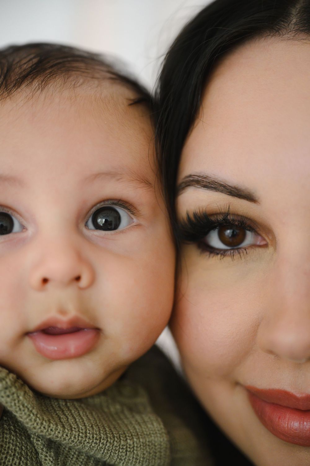 Close-up portrait of wide-eyed baby and dark-haired mother with glam makeup cheek to cheek.