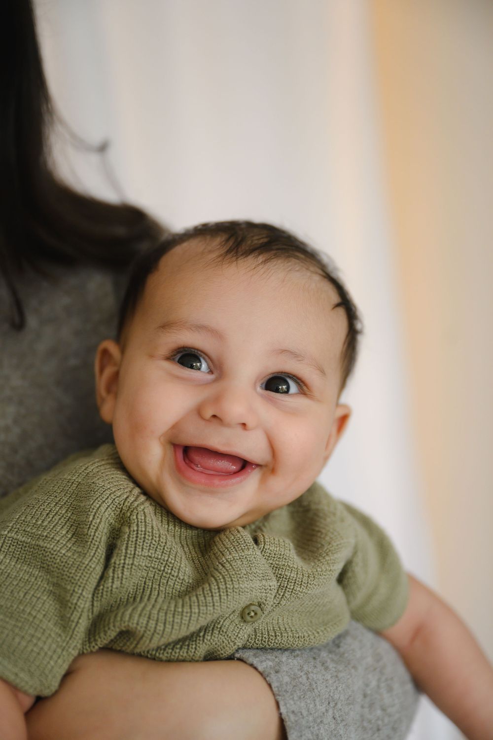 Happy smiling baby in sage green knit romper being held, showing big gummy smile.