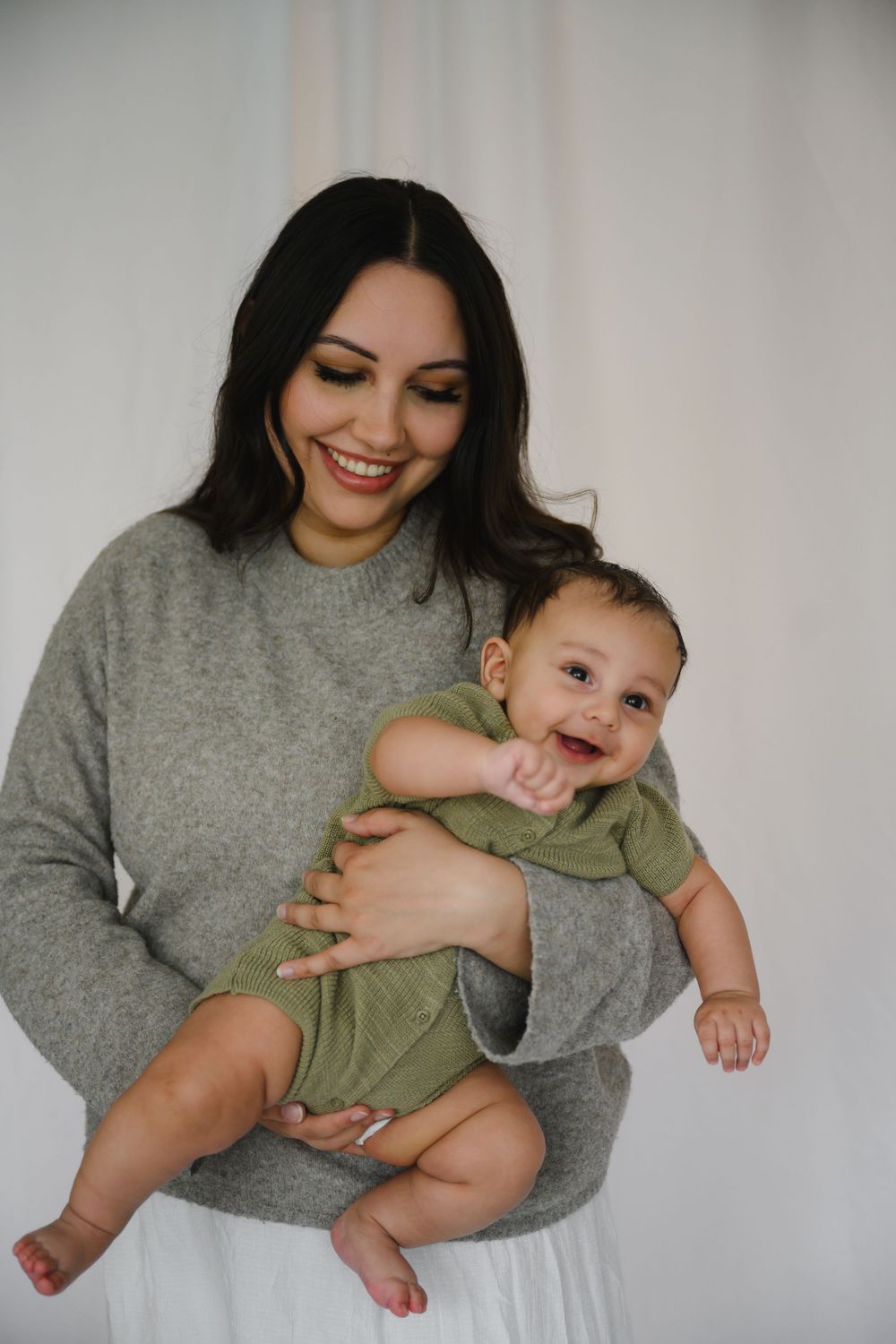 Joyful dark-haired mother in gray sweater cradling cheerful baby in green romper indoors.