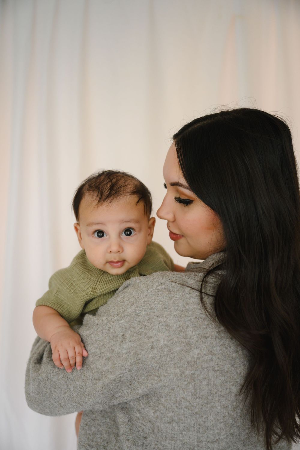 Dark-haired mother in gray sweater holding baby in green knit outfit against white backdrop.