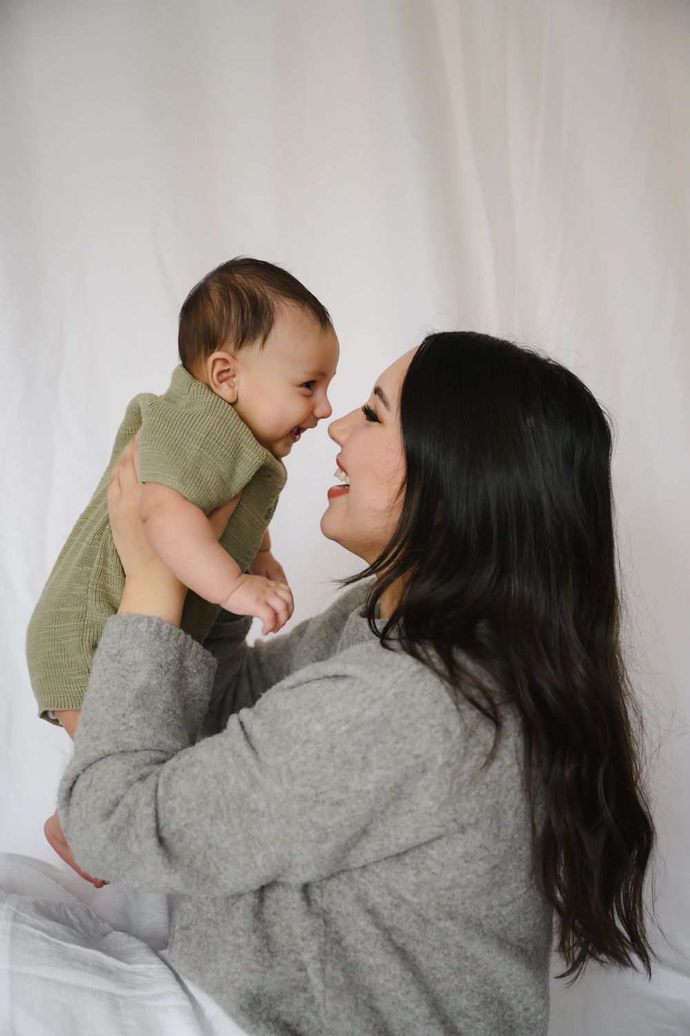 Loving mother lifting smiling baby in green romper for an eskimo kiss against white background.