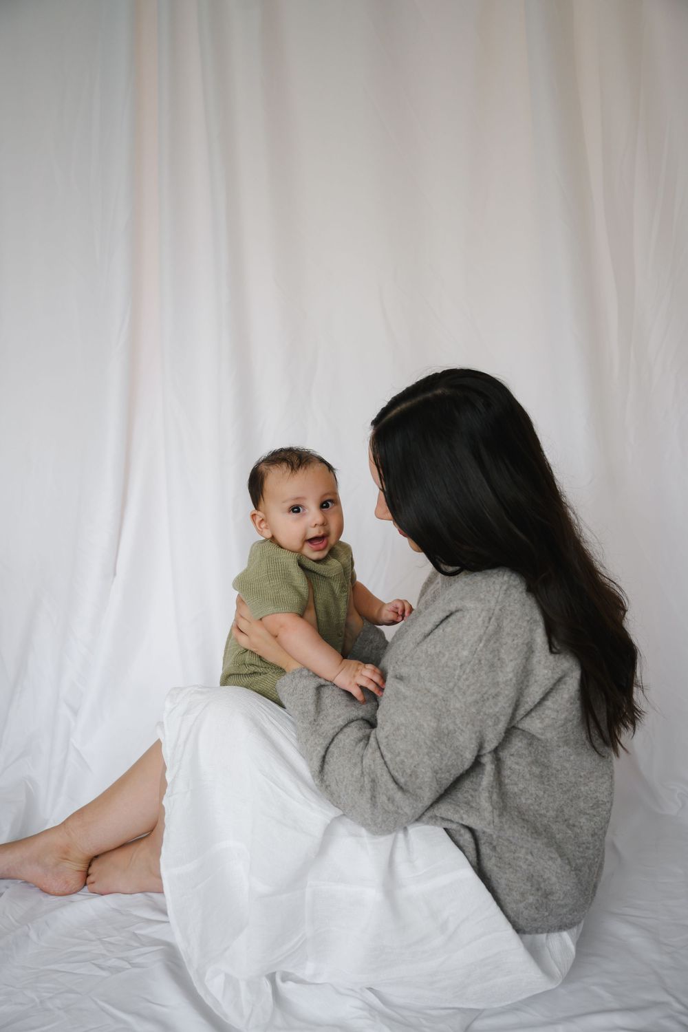 Mother in gray sweater sitting on white fabric floor holding happy baby in green romper.