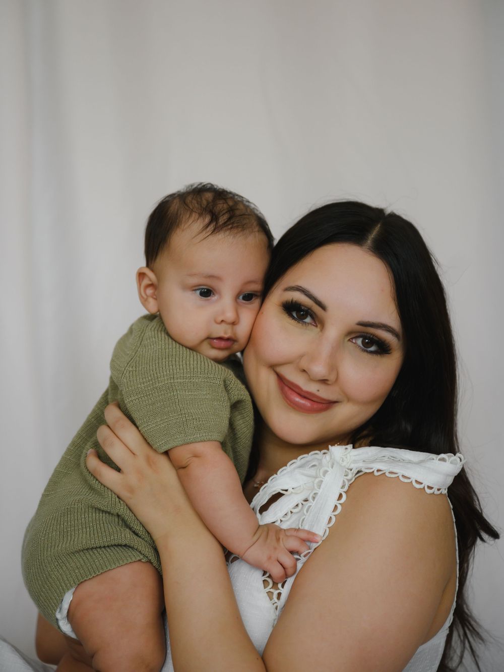 Smiling dark-haired mother in white top cuddling baby in green knit romper against white backdrop.