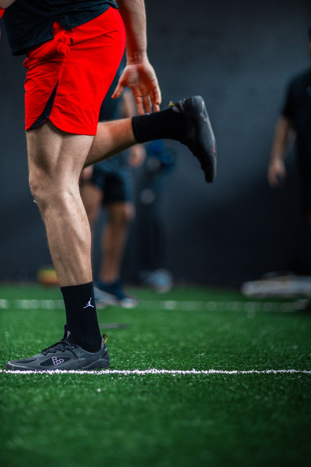 Athlete in red shorts and Jordan sneakers performing high-knee drills on an indoor artificial turf training facility.