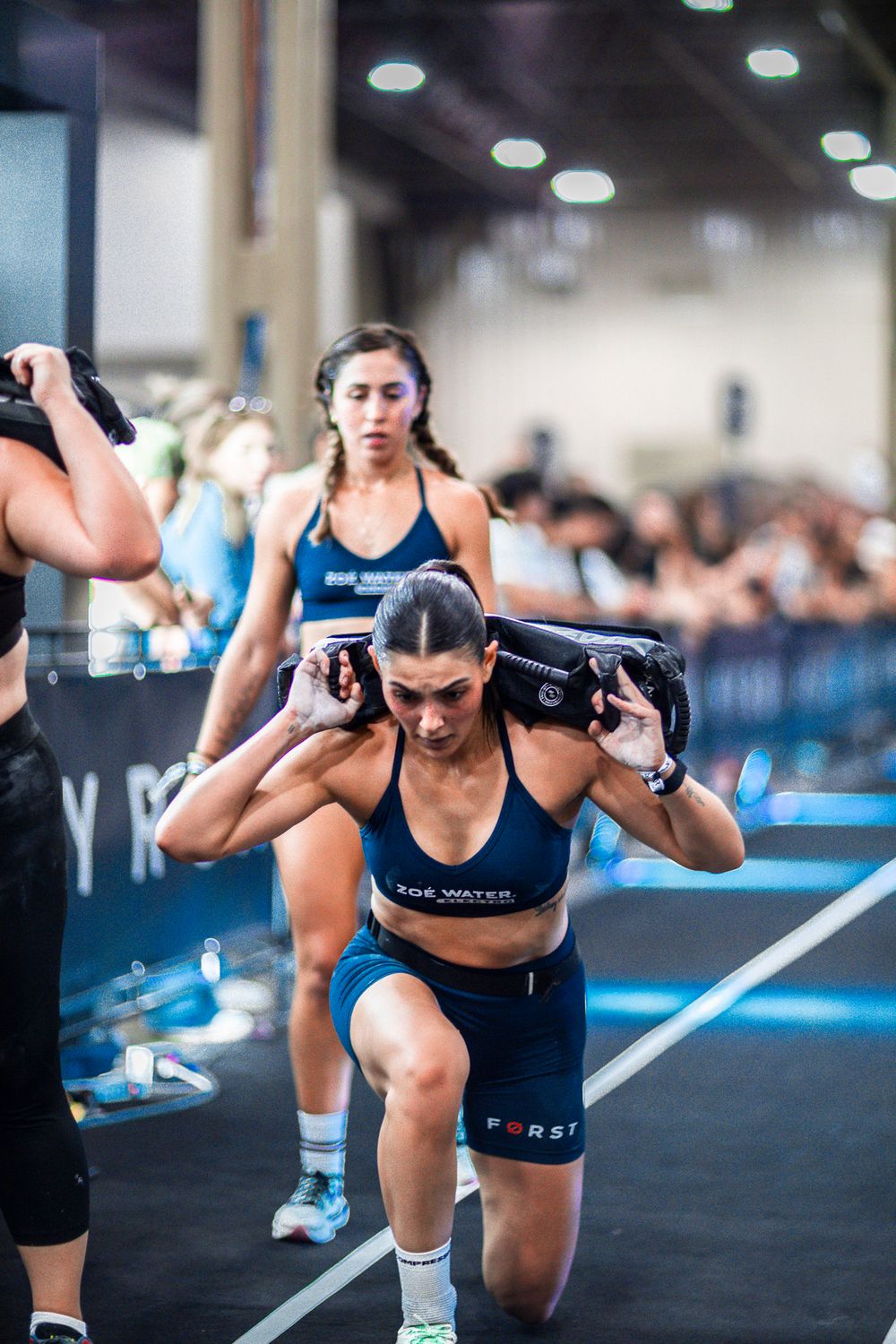 Athletes compete in an intense CrossFit competition carrying heavy sandbags across a gym floor during a fitness event.