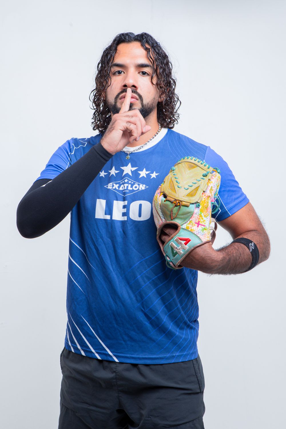 Baseball player in blue LEO Exatlon jersey holds glove, making silence gesture against white background.