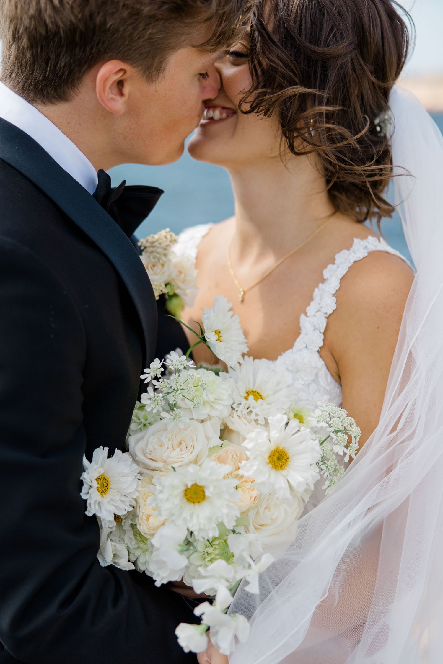 A romantic wedding moment with a white bouquet of roses and daisies held between the couple sharing a kiss.