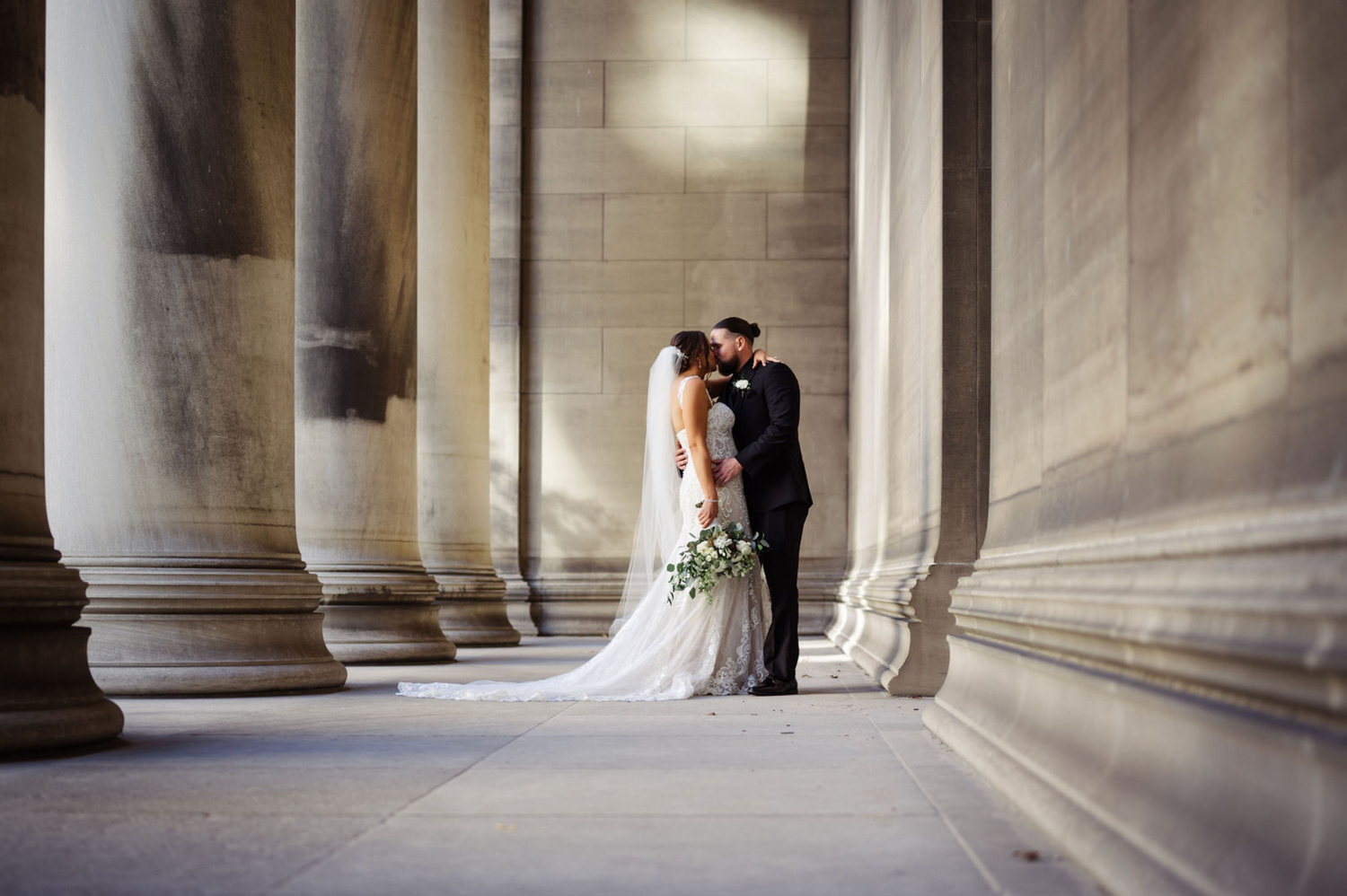 Bride and groom share a kiss between grand stone columns in a stunning wedding portrait.