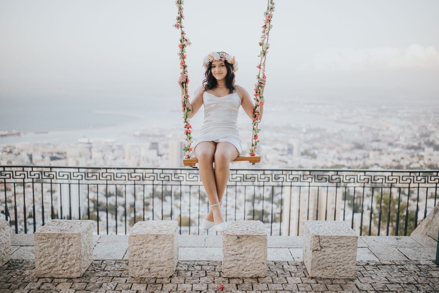 Person in white dress sitting on decorative swing overlooking city skyline.