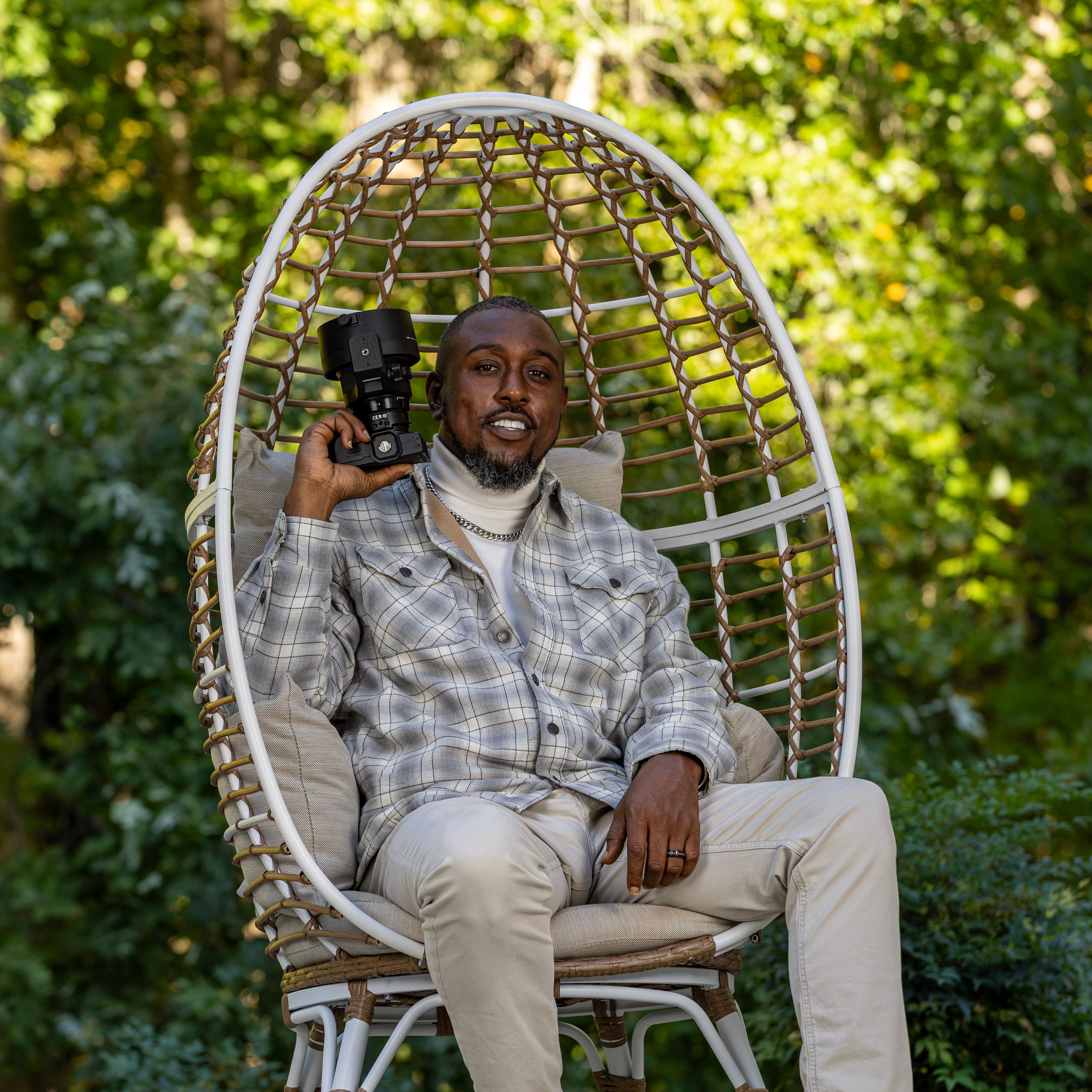 Someone relaxes in a white woven hanging chair outdoors surrounded by lush greenery. | Shot By Lo™