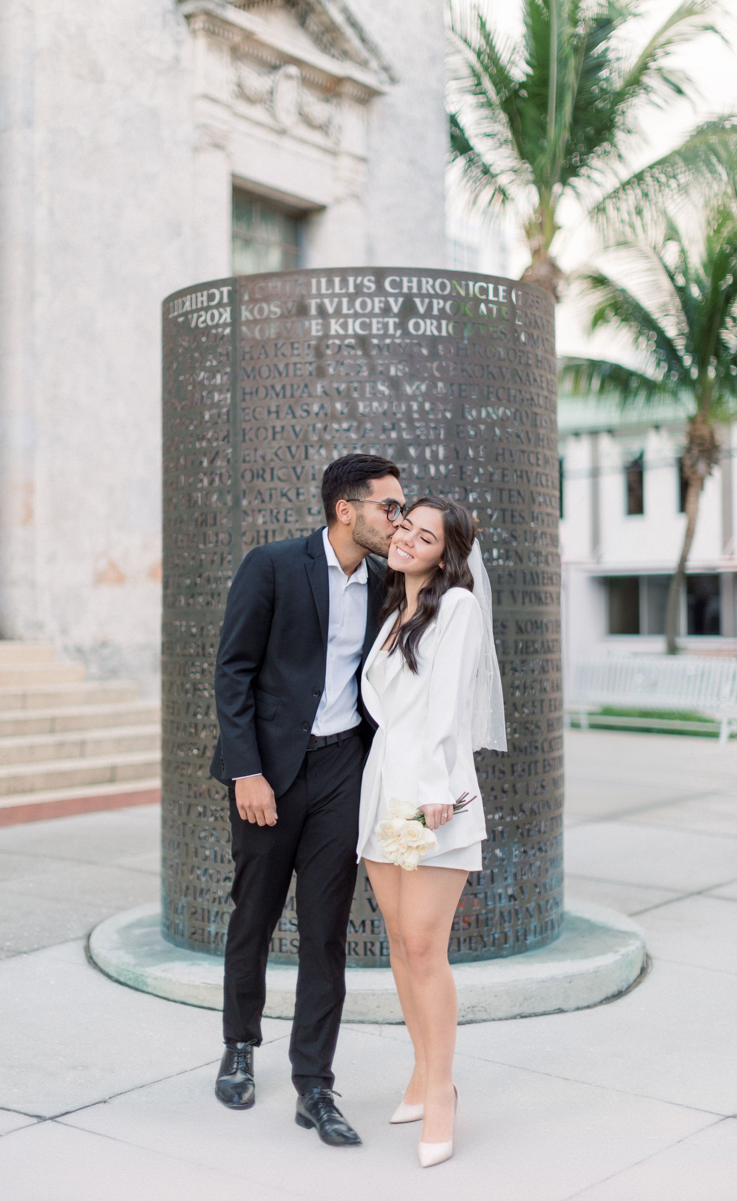 A couple shares romantic moment by engraved stone pillar near palm tree.