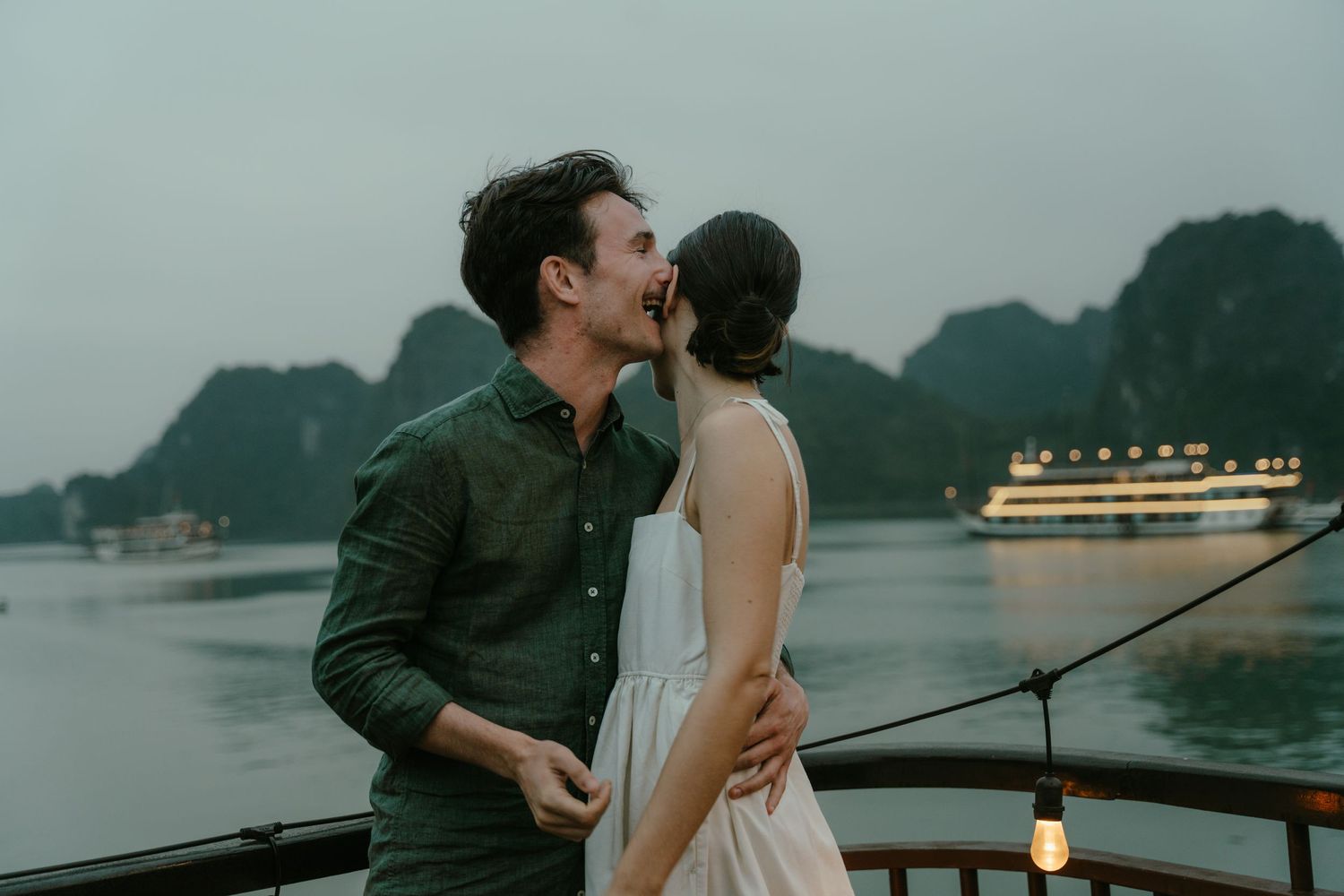 A romantic couple shares an intimate moment on a boat deck with limestone mountains and a cruise ship in the background.
