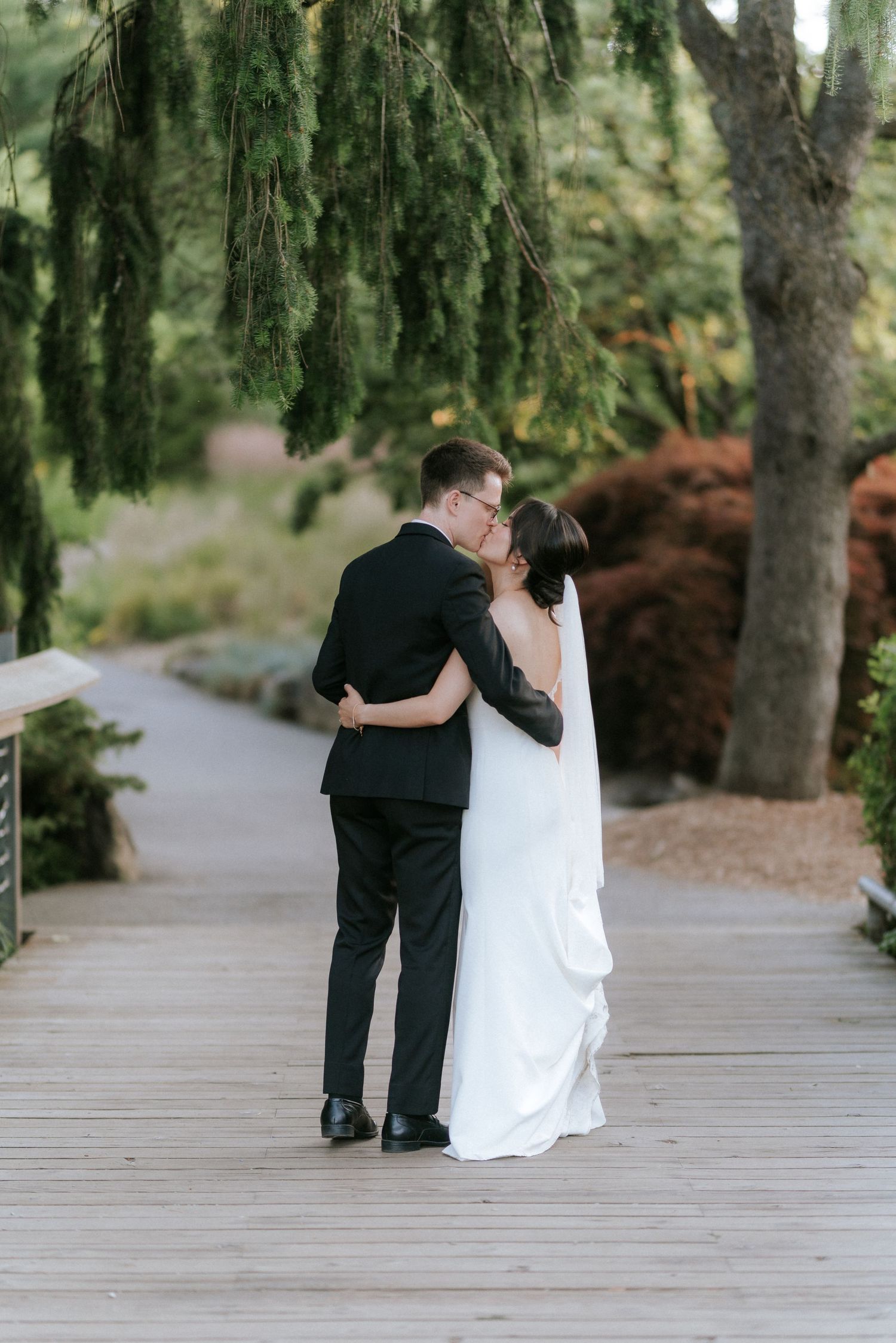 A romantic couple in wedding attire share an intimate moment together on a wooden boardwalk surrounded by trees.