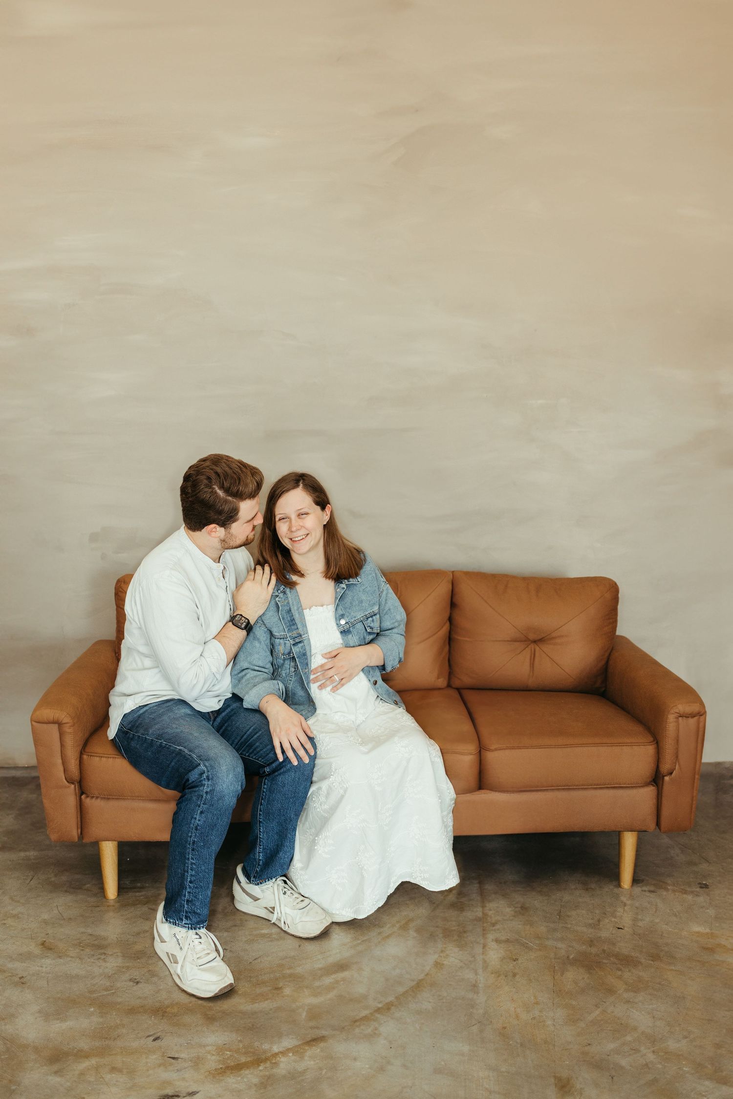 Happy couple sitting on brown leather sofa, pregnant woman in white dress and denim jacket with partner.