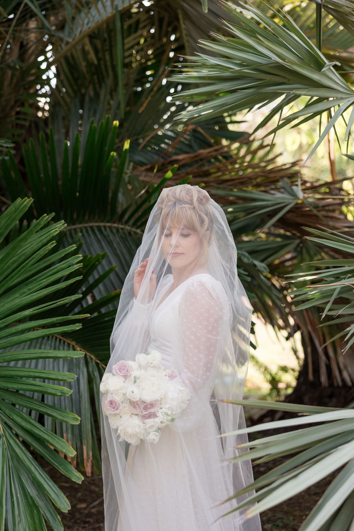 A bride in a delicate lace dress and veil poses among tropical palm fronds in a garden setting.