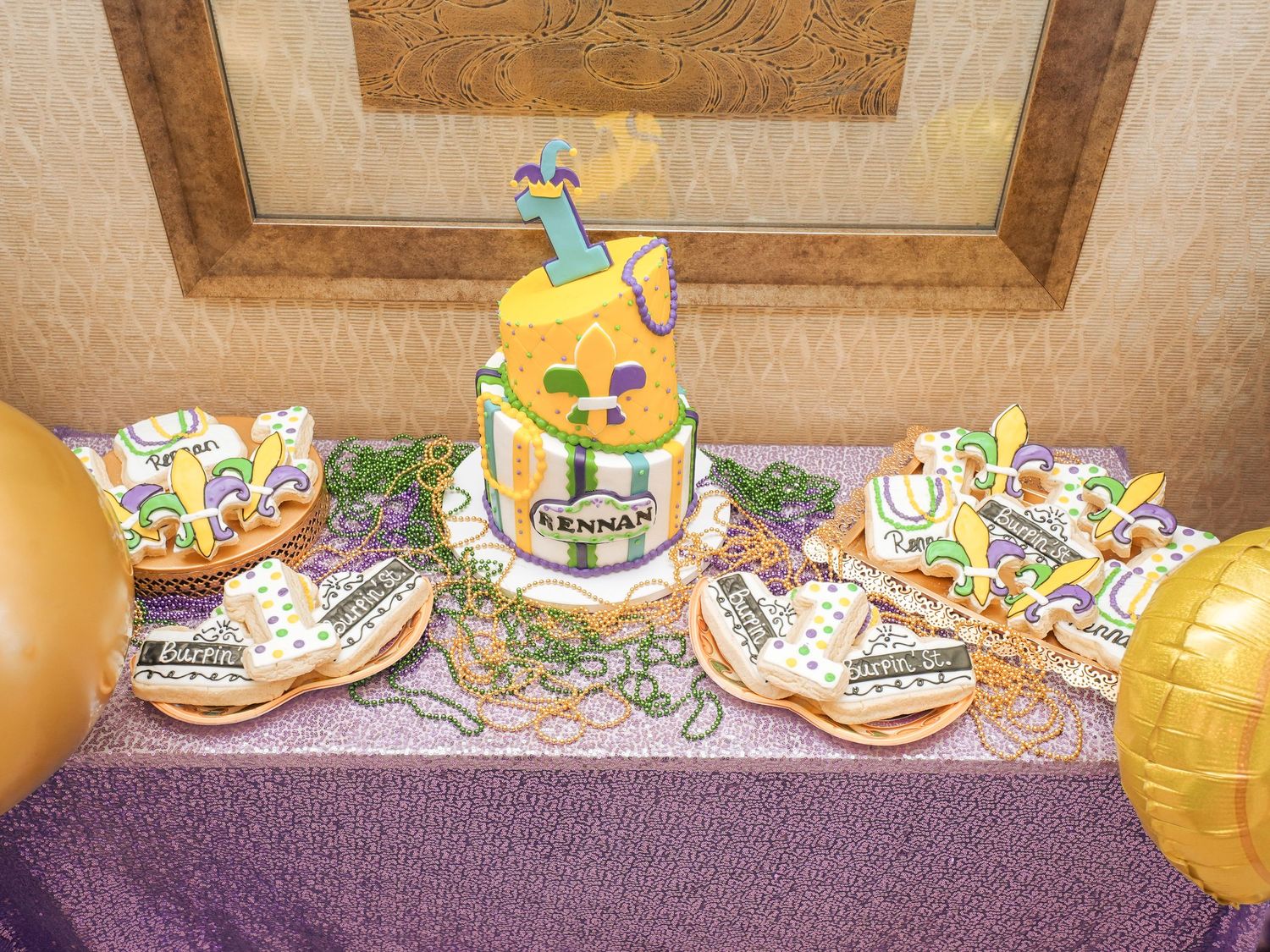 Festive dessert table with colorful birthday cake and decorated cookies on purple tablecloth.