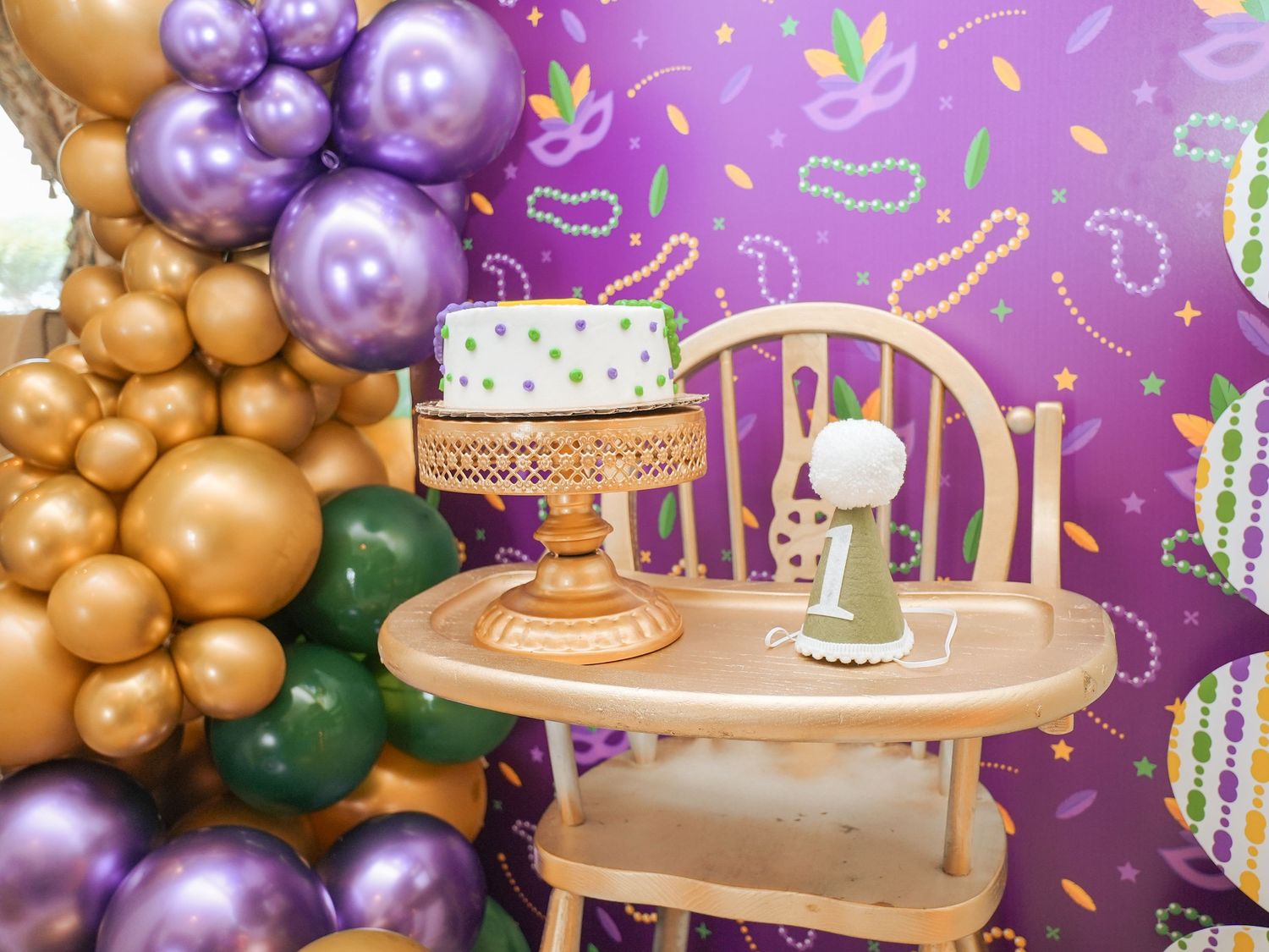 Birthday party setup with gold and purple balloon arch, wooden chair and decorated table.