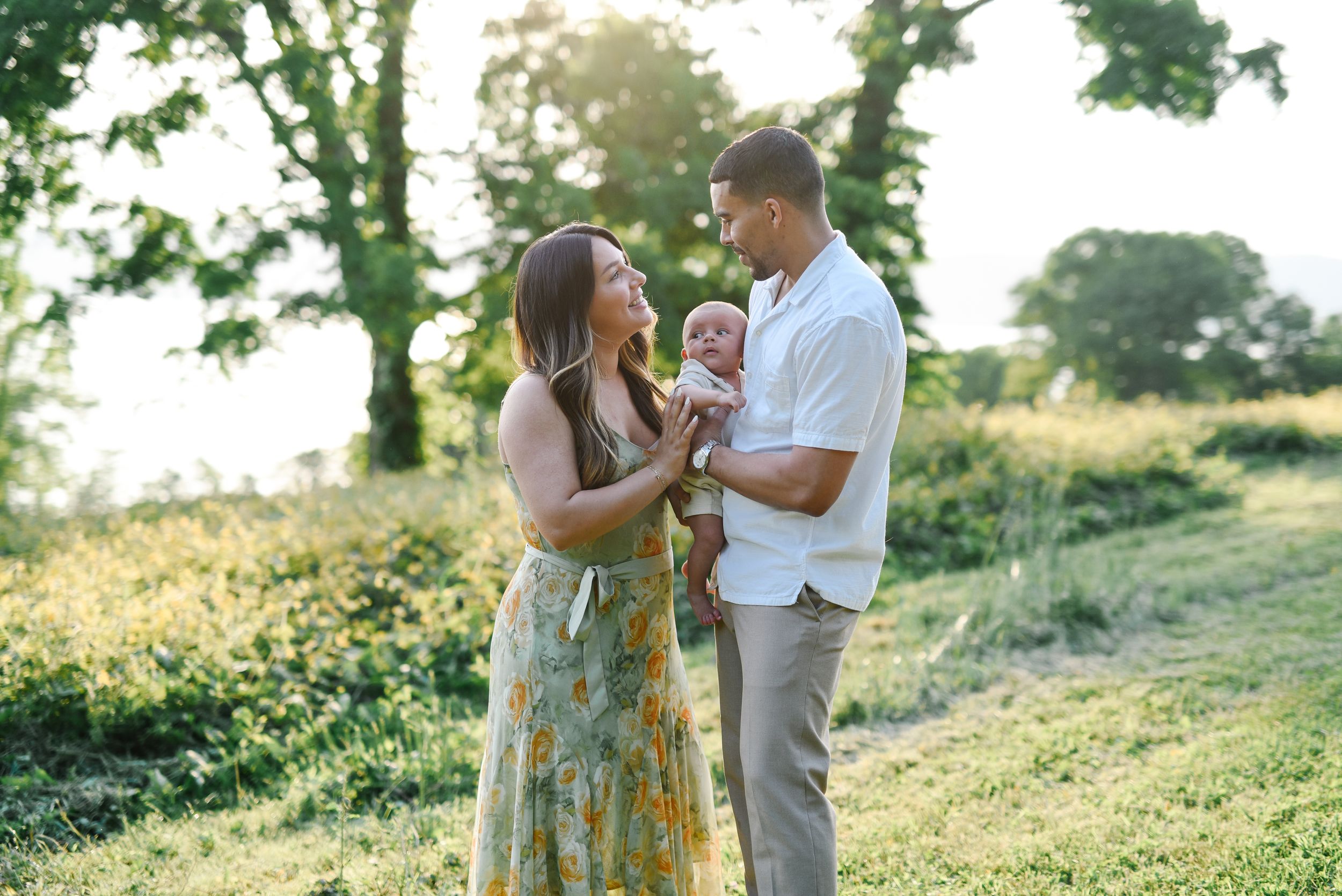 Couple shares intimate moment with baby on grassy hillside at sunset.