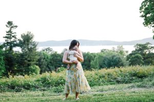 Woman in floral dress holds baby while standing on hilltop overlooking scenic view.