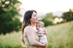 Mother in floral dress holds smiling baby against sunset sky.