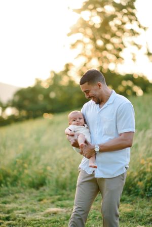 Father in white shirt cradles baby while standing in golden field at sunset.