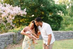 A couple holds their baby while standing near a stone wall with blooming flowers in the background.