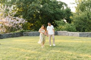 A couple walks together on grass near a stone wall during golden hour.