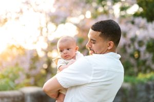 A person in white holds a baby while standing against a sunset-lit garden backdrop.