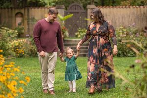 Family holding hands and walking together in a blooming garden with yellow flowers and a wooden fence in background.