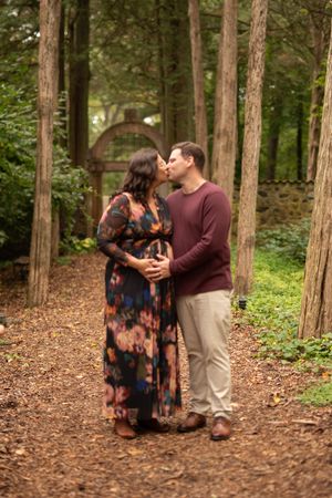 A couple embraces intimately among tall pine trees on a forest floor covered in brown needles.