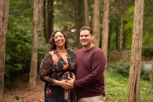 A couple stands together smiling in a wooded forest setting wearing fall-colored clothing.