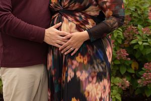 A close-up of hands embracing a pregnant belly, wearing a colorful floral dress against a green garden background.