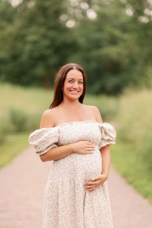 Expectant mother in off-shoulder cream dress stands on path surrounded by greenery.