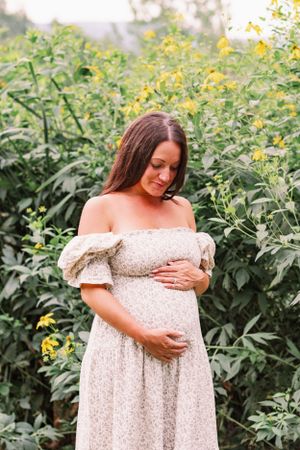 Expectant mother in cream dress poses among yellow wildflowers and greenery.