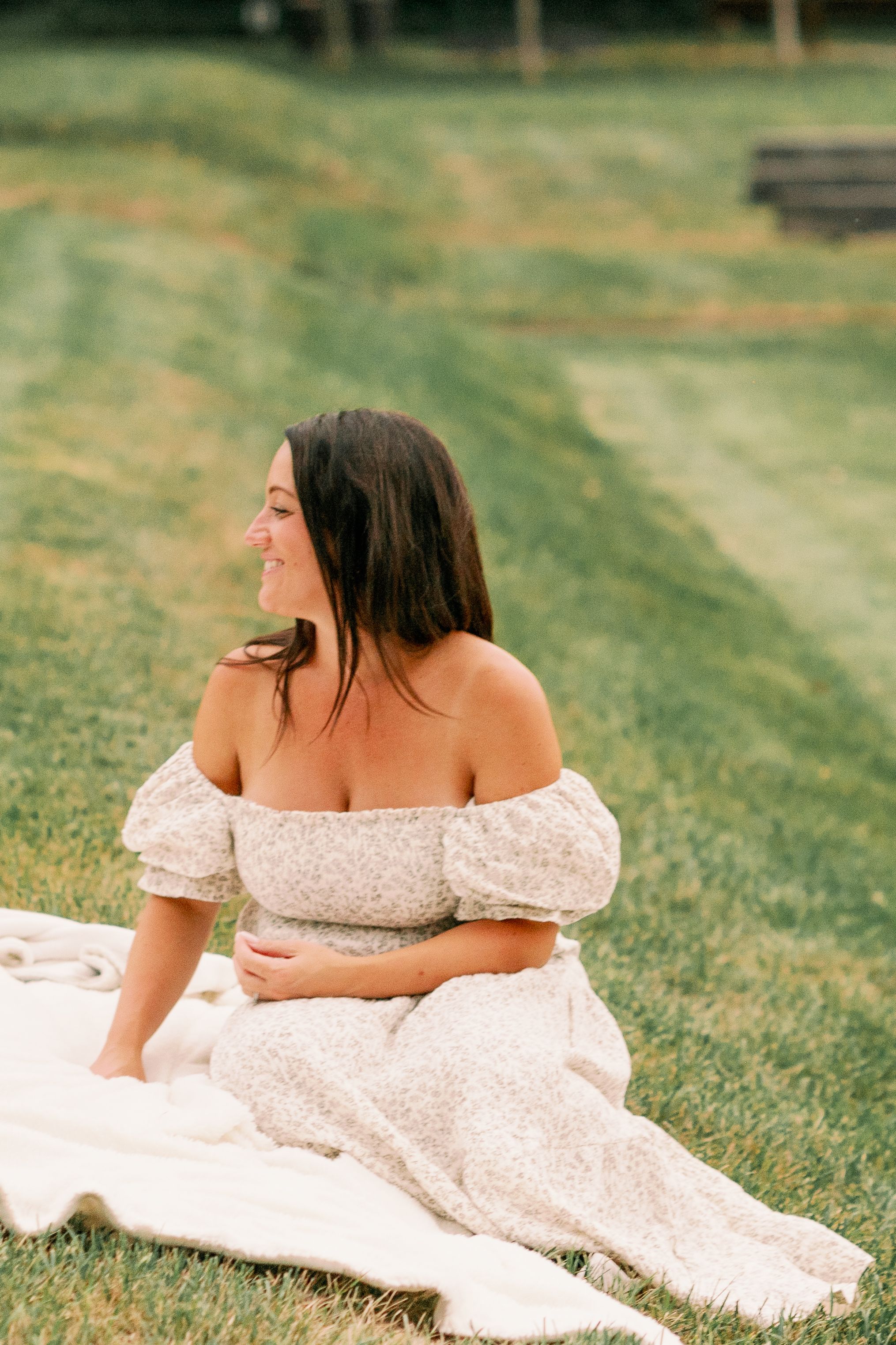 Woman in off-shoulder dress sits on blanket in grassy field during summer.