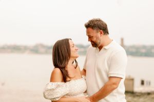 Couple embraces near waterfront at sunset wearing white and cream colored outfits.