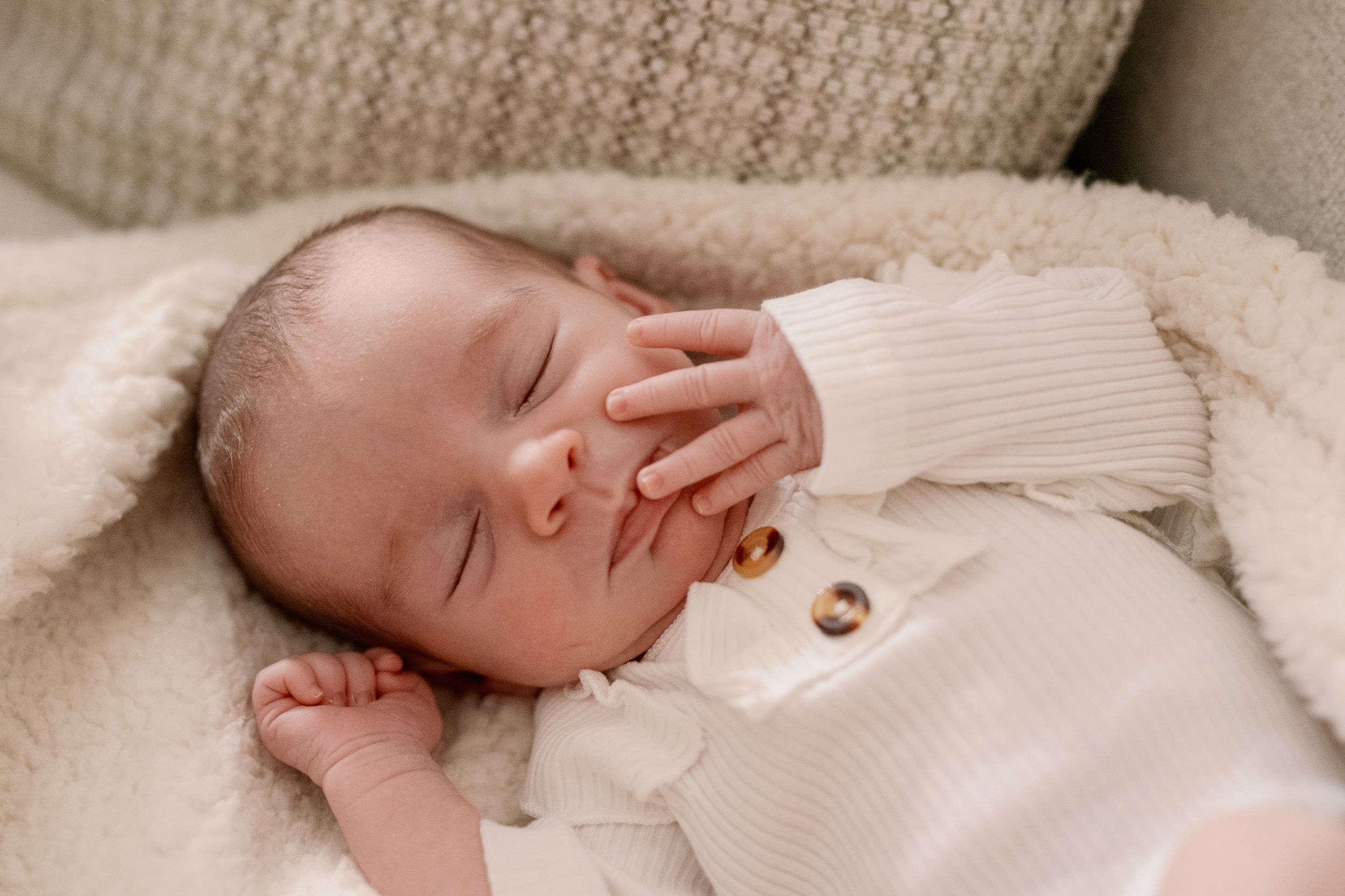 Sleeping infant in white knit clothing rests peacefully on cream blanket.