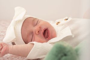 Newborn baby in white outfit yawning while lying on pink fabric.
