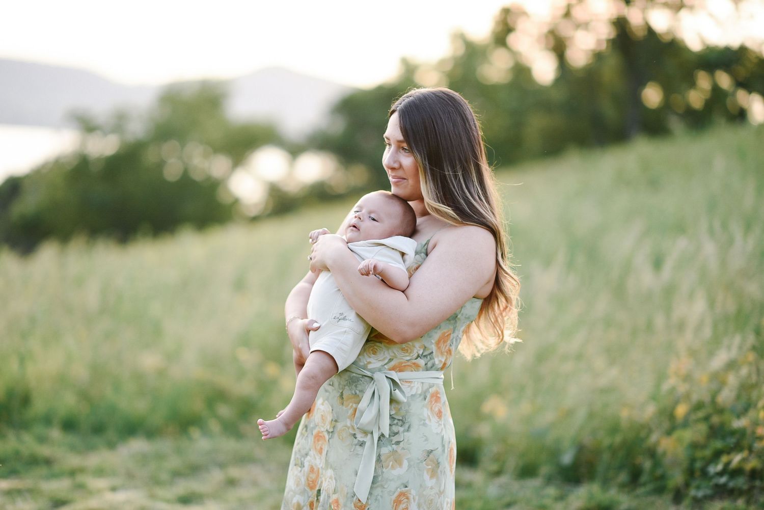 A person in floral dress holds infant while standing in grassy field at sunset.