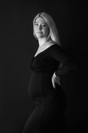 A black and white maternity portrait of a person in a long sleeve dress posing against a dark background.