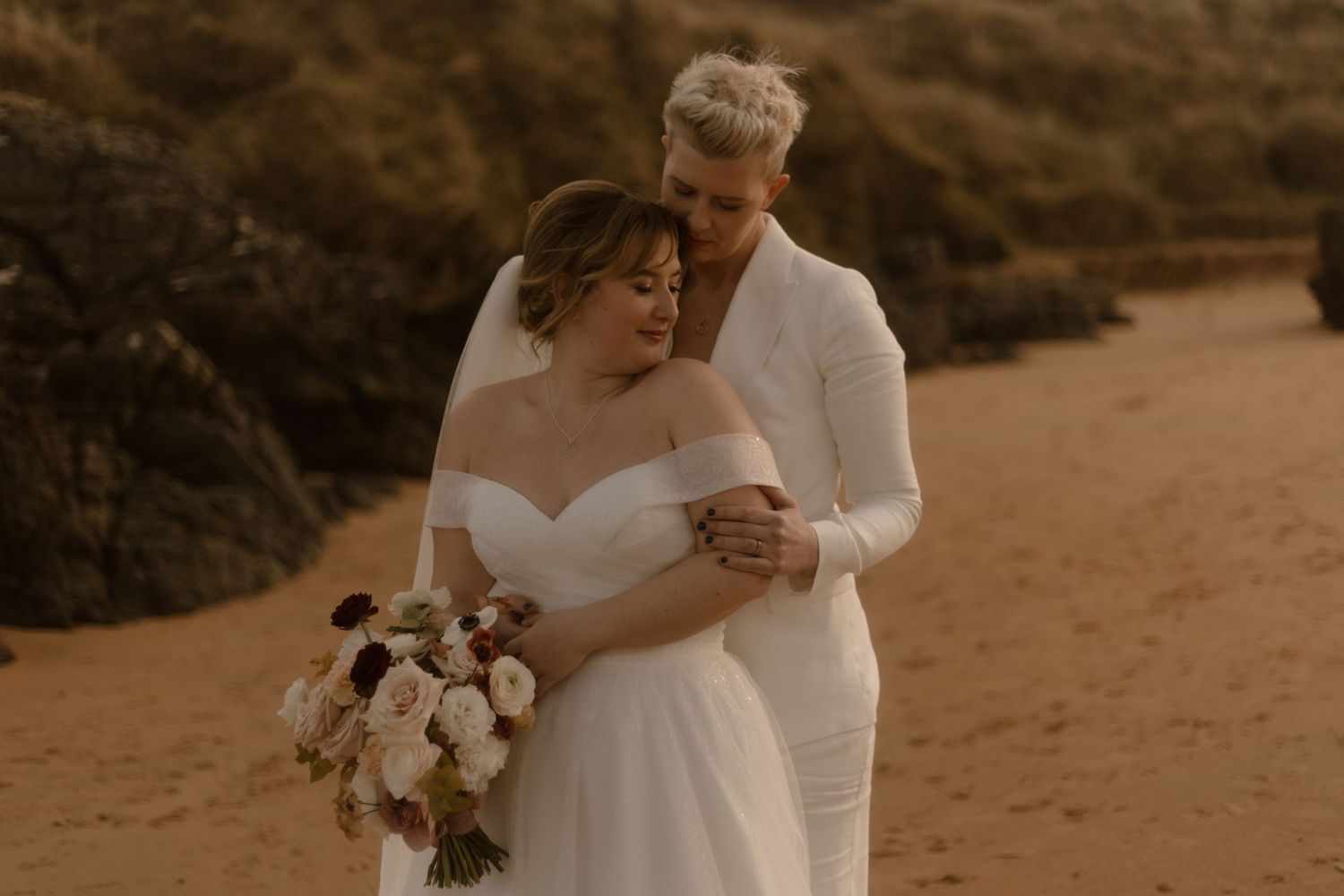 Intimate beach wedding moment with couple embracing against golden sunlit cliffs.