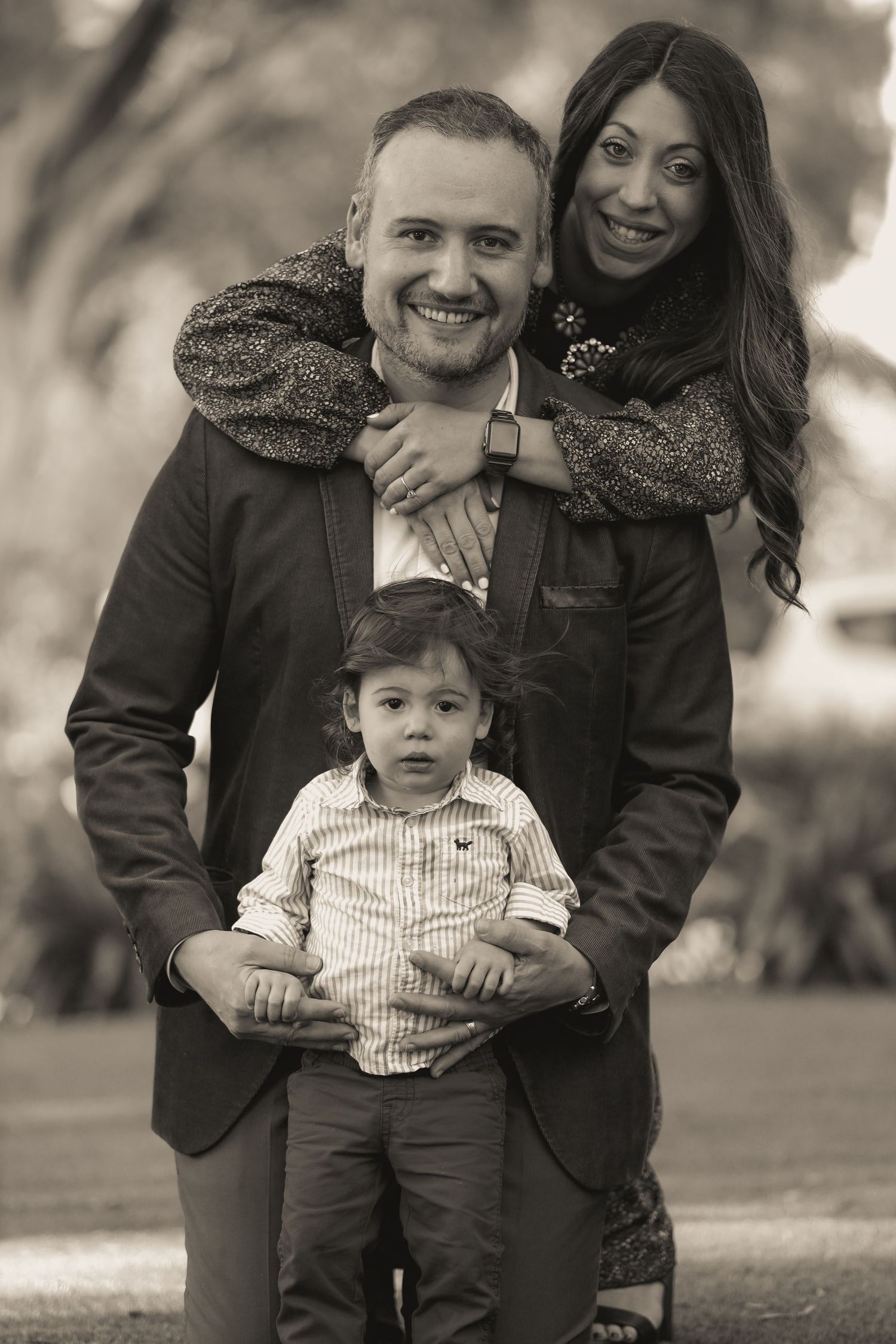 Black and white portrait of smiling couple with young child in park.
